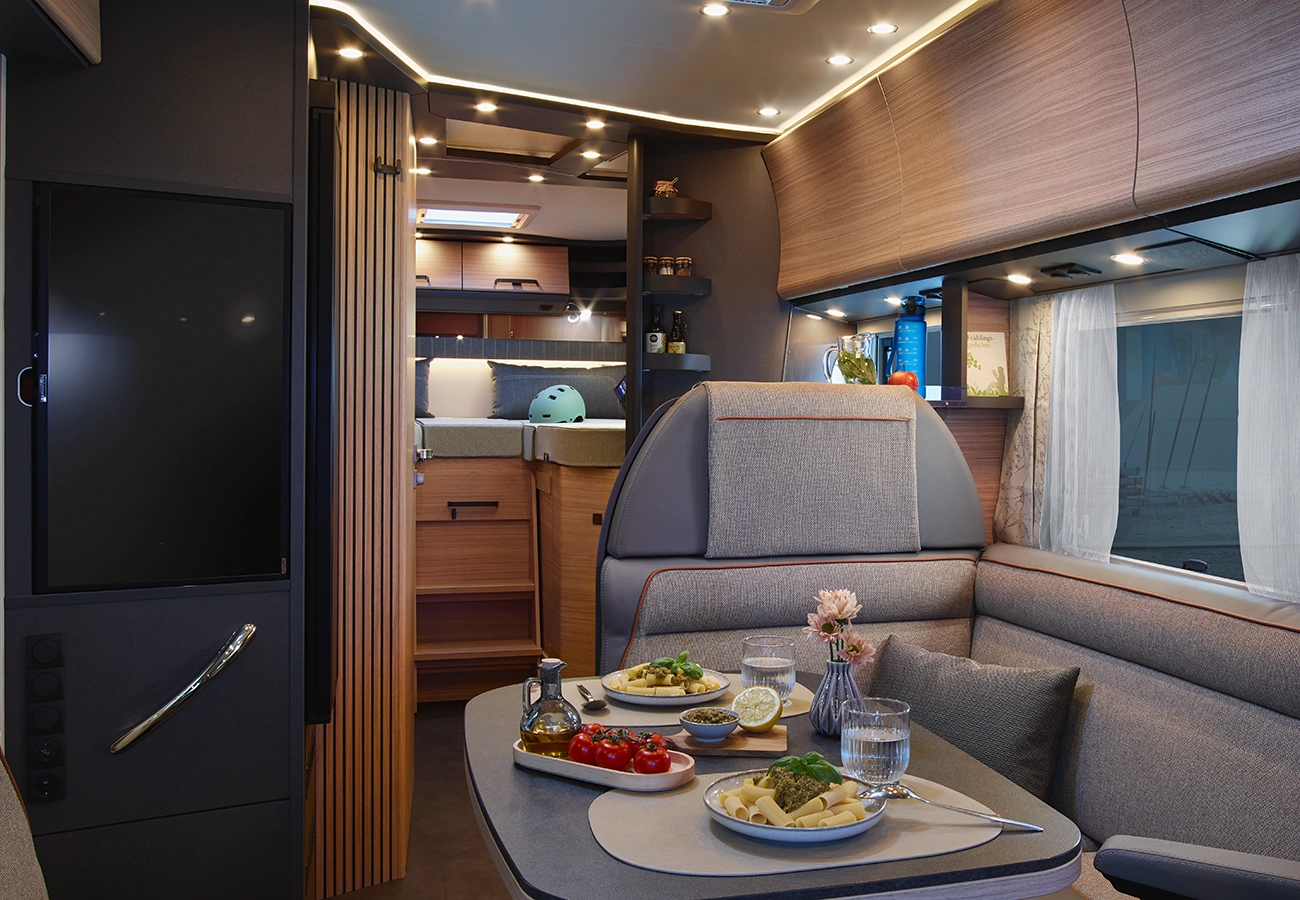 Ein modernes Wohnmobil-Interieur mit einem Esstisch für zwei Personen, an dem Nudeln, Salat und Gemüse aufgetischt werden. Der Raum ist gut beleuchtet, mit gemütlichen Sitzgelegenheiten, Holzakzenten und einem ordentlich gemachten Bett im Hintergrund.