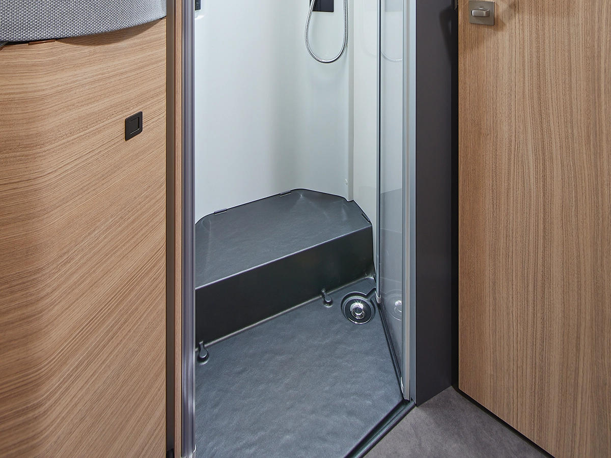 Eine kompakte Duschkabine mit Glastür und grauem, strukturiertem Boden in einem modernen Wohnmobil oder Camper, umgeben von hellen Holzverkleidungen und Schränken.