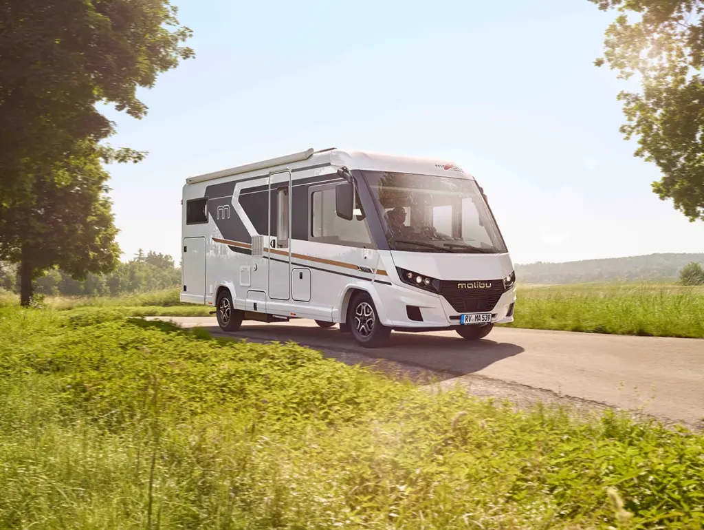 Ein weißes Wohnmobil fährt an einem sonnigen Tag eine Landstraße entlang, umgeben von grünem Gras und Bäumen, mit einem klaren Himmel im Hintergrund.