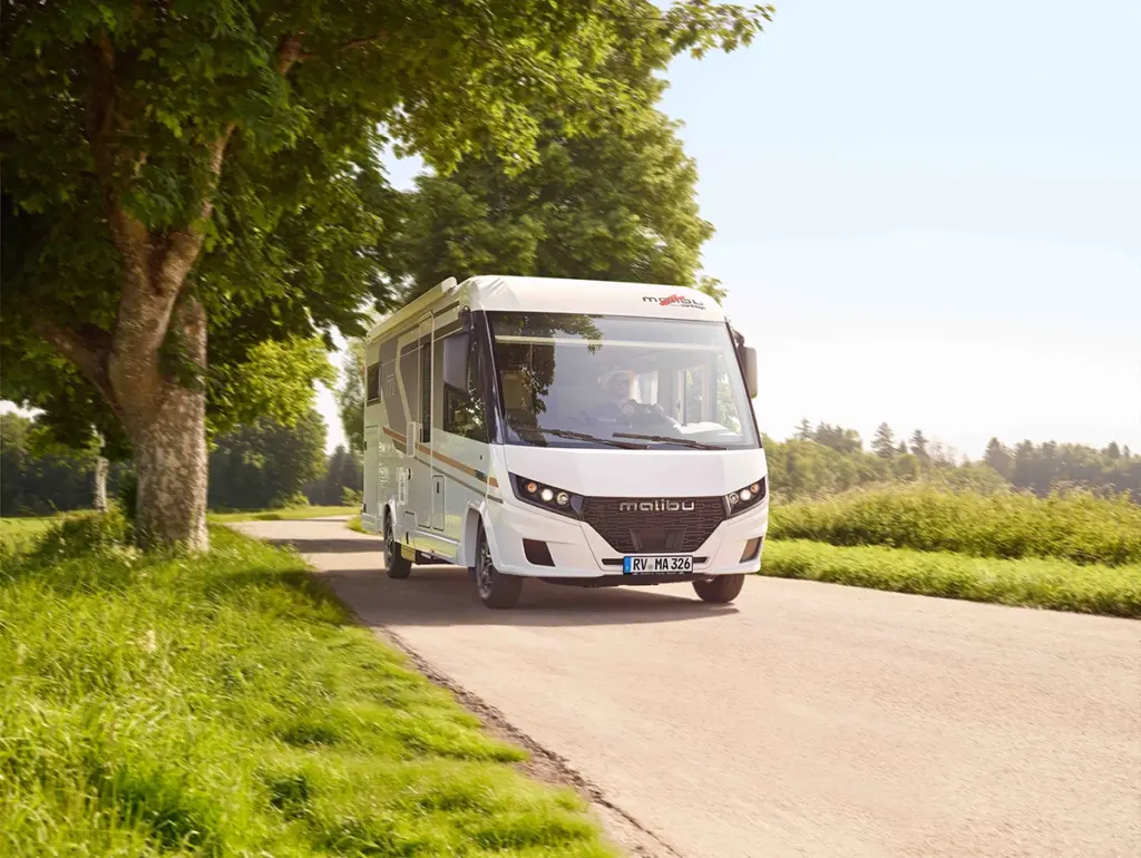 Ein weißes Malibu-Wohnmobil fährt auf einer Landstraße, die von grünem Gras und Bäumen gesäumt ist, unter einem klaren Himmel, wobei das Sonnenlicht durch die Blätter fällt.