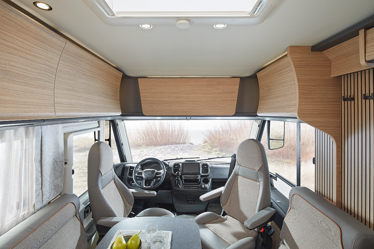 Innenraum eines modernen Wohnmobils mit zwei drehbaren Kapitänssesseln an der Vorderseite, einem zentralen Tisch mit Birnen und Gläsern, großen Fenstern und hellen Holzschränken, die eine helle, gemütliche Atmosphäre schaffen.