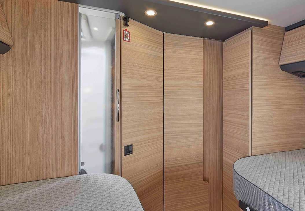 Modernes Wohnmobil-Interieur mit heller Holzvertäfelung, zwei grau gepolsterten Schlafbereichen, Hängeschränken und Deckeneinbauleuchten. Der Raum ist sauber, kompakt und effizient gestaltet.