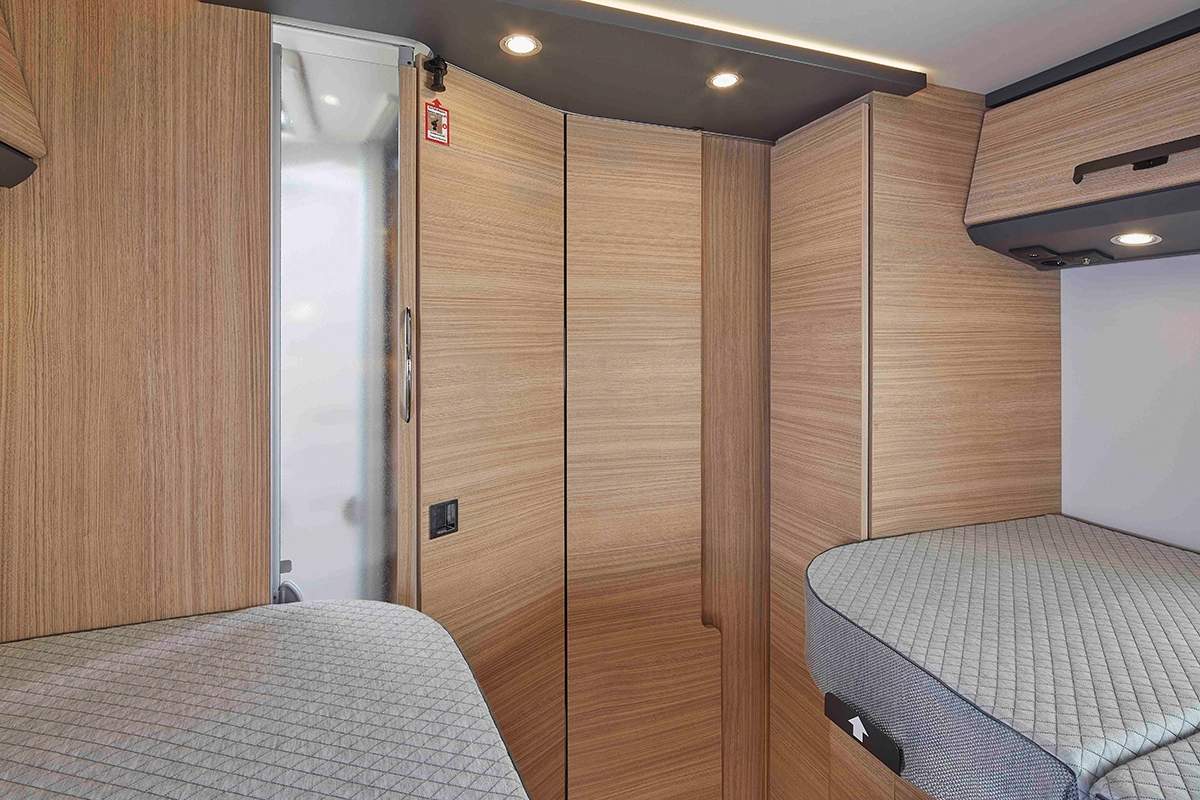 Modernes Wohnmobil-Interieur mit heller Holzvertäfelung, zwei grau gepolsterten Schlafbereichen, Hängeschränken und Deckeneinbauleuchten. Der Raum ist sauber, kompakt und effizient gestaltet.