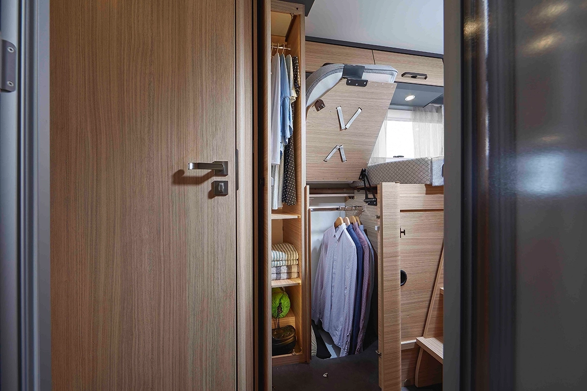 Ein moderner hölzerner Kleiderschrank mit offenen Türen, der hängende Hemden, gefaltete Kleidung, einen Korb und einige Ablagefächer zeigt. Natürliches Licht kommt von einem Fenster im Hintergrund.