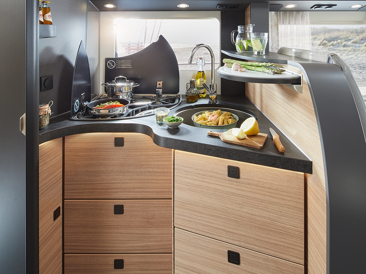 Moderne Wohnmobilküche mit Holzschränken, einem Herd, auf dem Gemüse gekocht wird, einer Spüle mit frischen Produkten, einem Schneidebrett mit Zitronen und einer kleinen Theke mit Salat und Getränken. Sonnenlicht strömt durch ein Fenster herein.