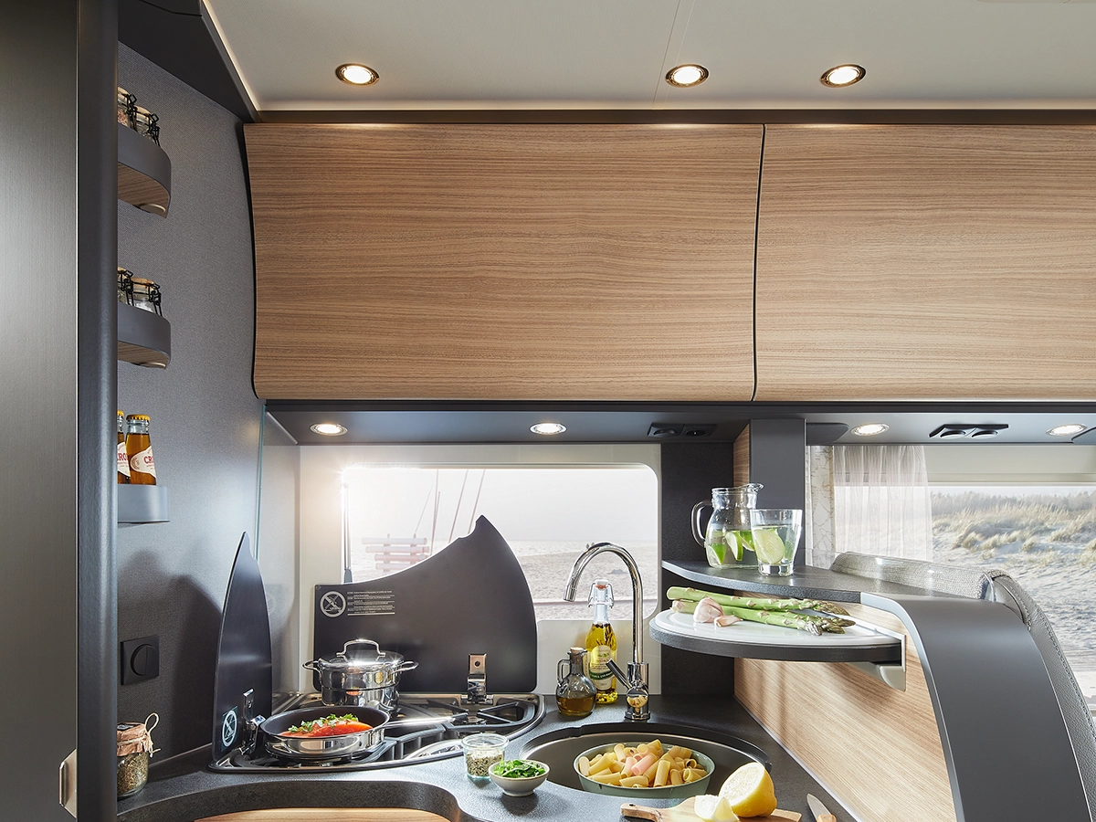 Moderne Wohnmobilküche mit Holzschränken, einem Herd mit Töpfen und Pfannen, einer Spüle, frischem Gemüse und Zitronen auf der Theke, Gläsern auf Regalen und einem Fenster mit Blick auf eine sandige Außenlandschaft.