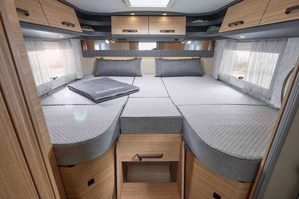 Das Innere eines modernen Wohnmobil-Schlafzimmers mit einem großen Bett mit grauer Bettwäsche, eingebauten Holzschränken und Schubladen, Fenstern mit transparenten Vorhängen und Stauraum über dem Kopf. Natürliches Licht durchflutet den gemütlichen, gut organisierten Raum.