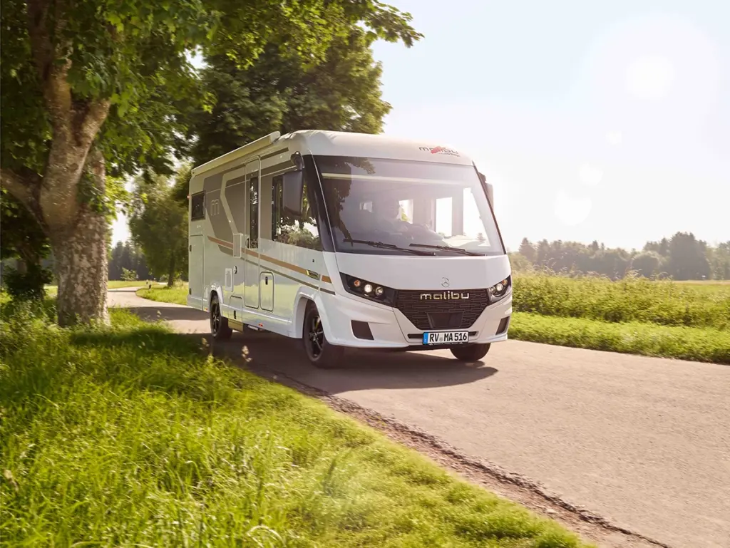 Ein weißes Malibu-Wohnmobil fährt auf einer schmalen Landstraße, die von grünem Gras und Bäumen gesäumt ist, unter einem strahlenden, sonnigen Himmel.