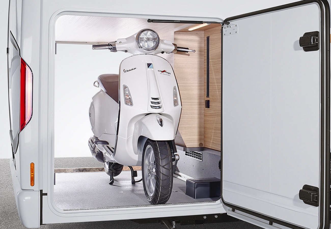 Ein weißer Vespa-Roller ist sicher im hinteren Stauraum eines Wohnmobils geparkt, wobei die Tür des Stauraums geöffnet ist und den Roller sowie einen Teil des Innenraums des Fahrzeugs freigibt.
