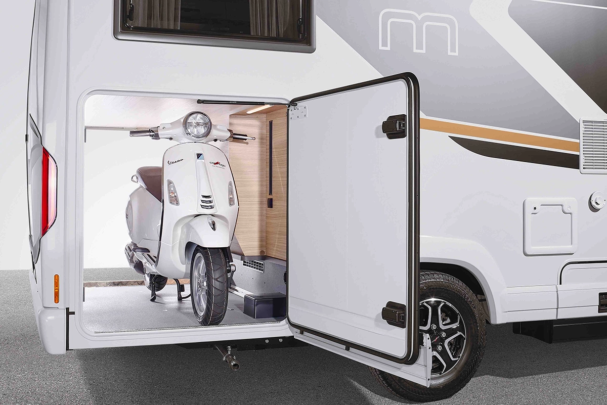 Ein weißer Motorroller ist sicher in einem Seitenfach eines weißen Wohnmobils verstaut, wobei die Tür des Fachs geöffnet ist und der Motorroller ordentlich darin verstaut ist.