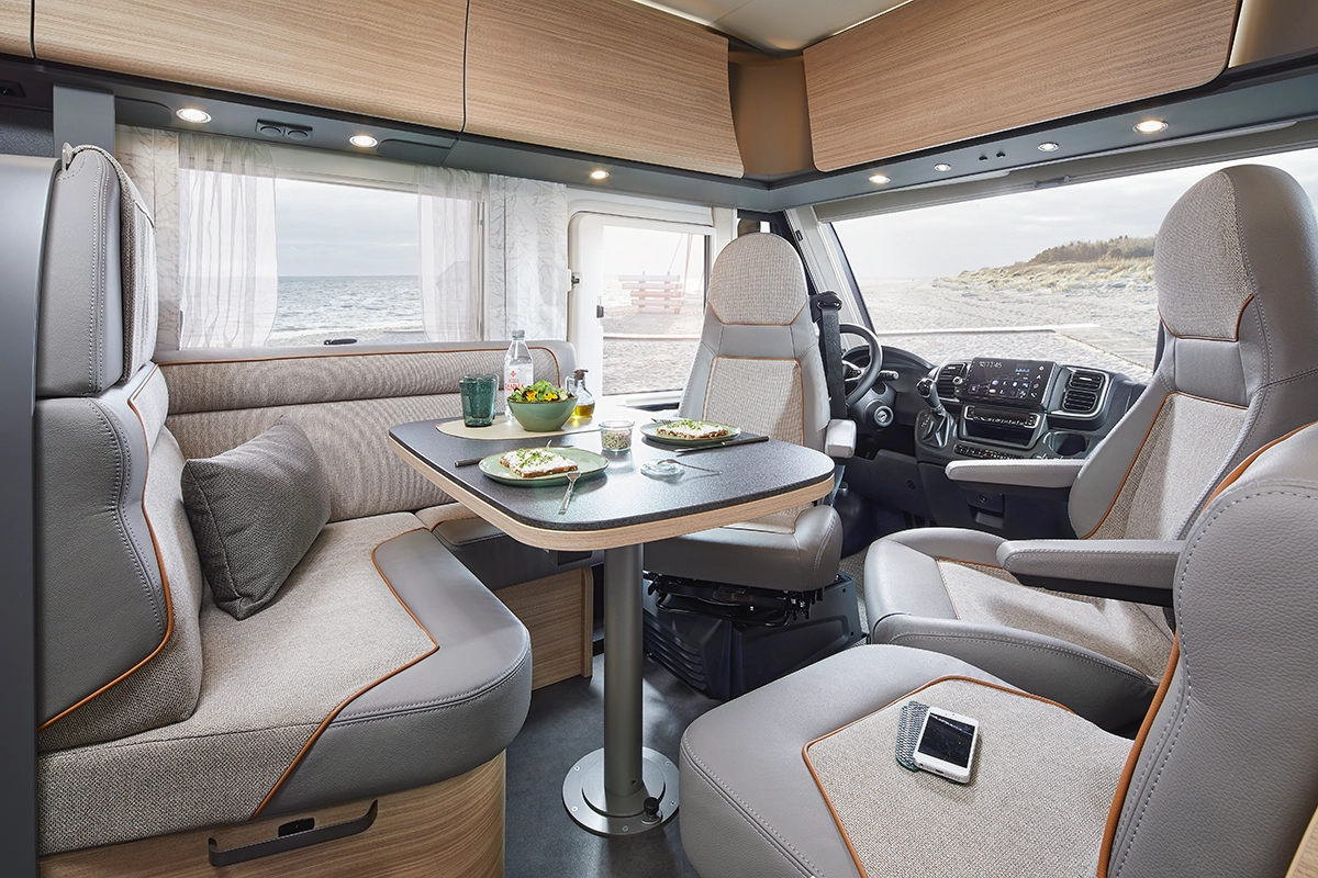 Das Innere eines modernen Wohnmobils zeigt einen Essbereich mit vier Sitzen um einen Tisch mit Salattellern, Gläsern und einer Flasche Wasser, der in der Nähe eines Strandes geparkt ist und durch große Fenster einen Blick aufs Meer bietet.