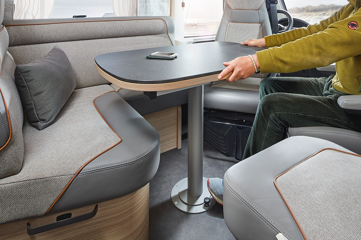 Eine Person, die in einem Wohnmobil sitzt, fährt einen klappbaren Tisch zwischen grauen gepolsterten Sitzen mit Holzakzenten aus. Auf dem Tisch liegt ein Smartphone, und durch die nahe gelegenen Fenster strömt Sonnenlicht.