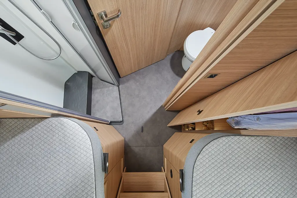 Blick von oben auf ein kompaktes Wohnmobil-Badezimmer mit einer Toilette, einer Dusche, Holzschränken und zwei Betten mit gestepptem grauem Bettzeug auf jeder Seite. In einem der Schränke ist ein Hemd zu sehen.