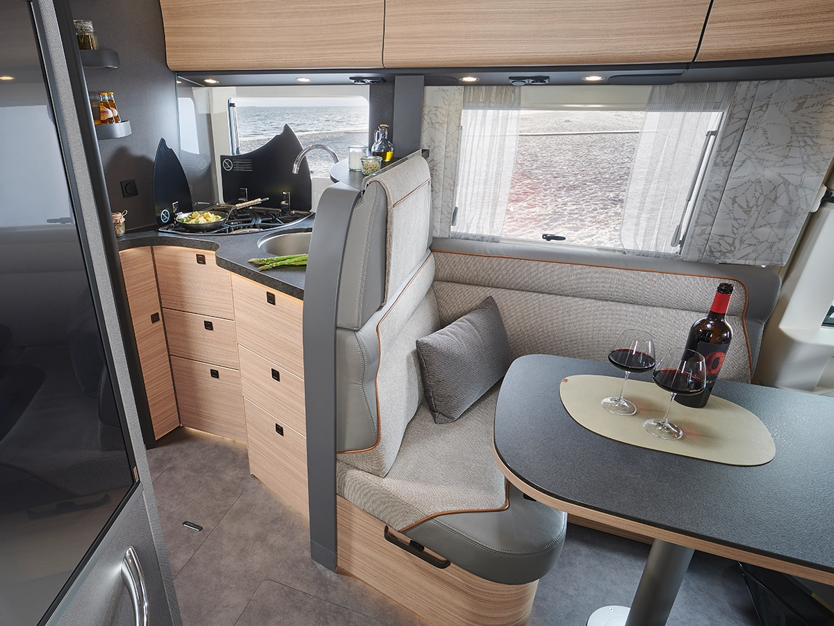 Modernes Wohnmobil-Interieur mit hellen Holzschränken, einer kleinen Küchenzeile, einer gepolsterten Essbank und einem Tisch mit zwei Gläsern Rotwein und einer Weinflasche neben einem großen Fenster mit Panoramablick.
