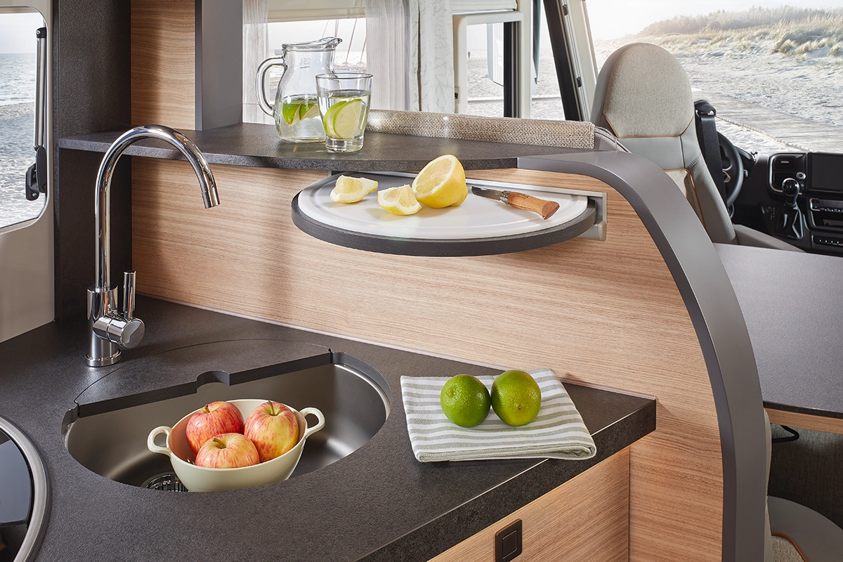 Eine moderne Wohnmobilküche mit einer runden Spüle mit Äpfeln, einem gestreiften Handtuch mit zwei Limetten, einem Schneidebrett mit Zitronenscheiben und einem Messer sowie einem Krug und einem Glas Zitronen-Limetten-Wasser auf dem Tresen neben einem Fenster mit Blick auf den Strand.