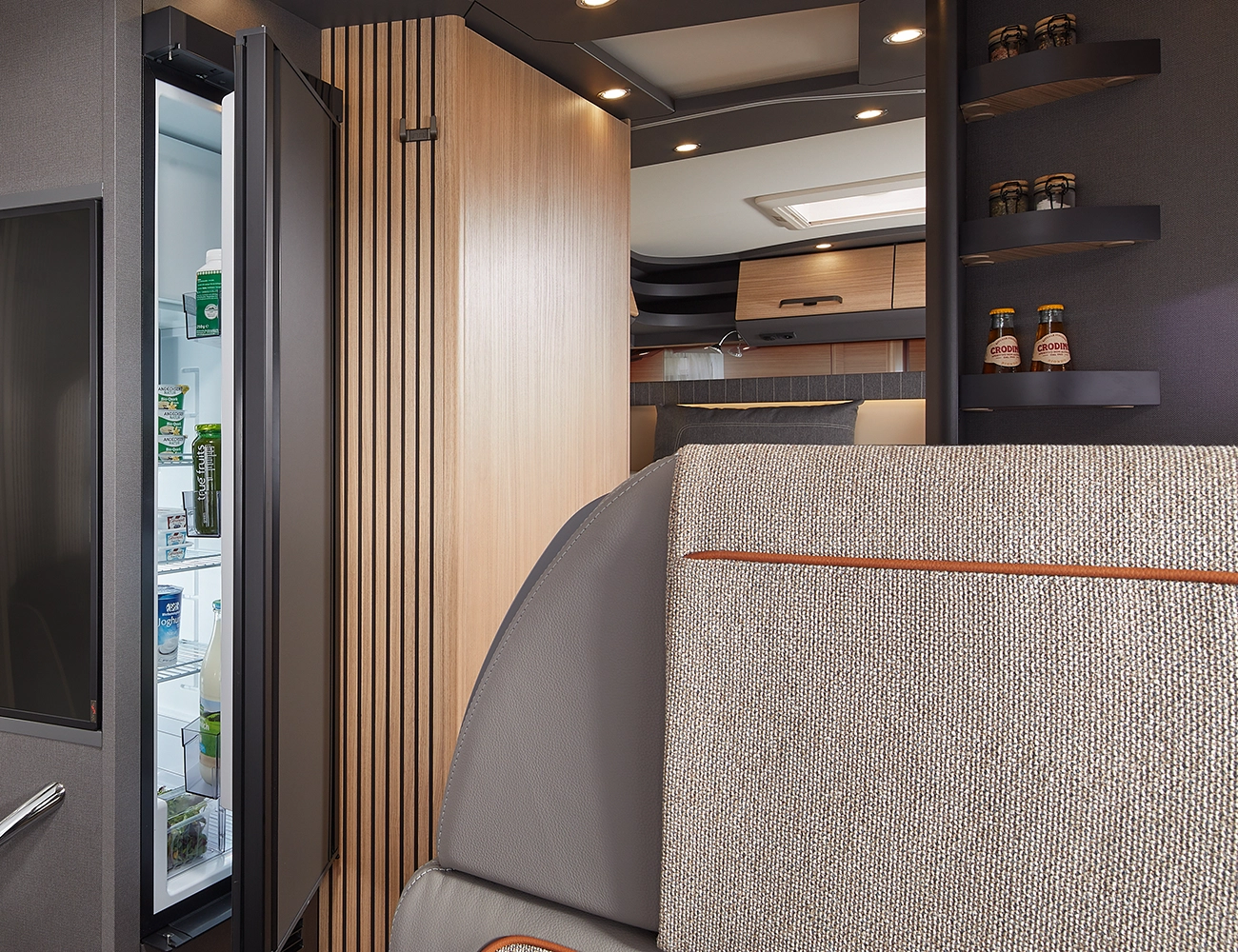 Modernes Wohnmobilinterieur mit einem offenen Kühlschrank mit Lebensmitteln, Holzschränken, Regalen mit Gläsern und Flaschen und einer Teilansicht eines gepolsterten Sitzes im Vordergrund.