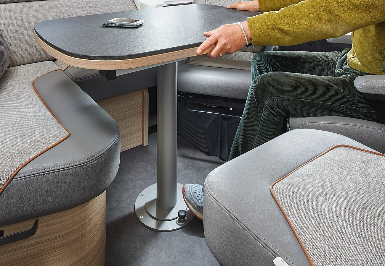 Eine Person in grüner Cordhose stellt einen modernen, schwarz umrandeten Tisch in einer gemütlichen Wohnmobil-Sitzecke mit grauen gepolsterten Bänken und einem Smartphone auf der Tischplatte ein.