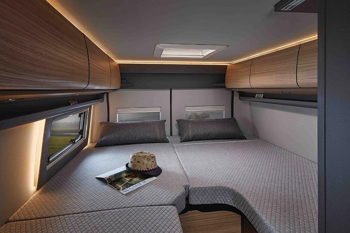 Interno di camper moderno e accogliente con letto matrimoniale con biancheria grigia, due cuscini, pensili in legno, due finestre, un cappello e una rivista posizionati sul letto sotto una luce d'ambiente soffusa.