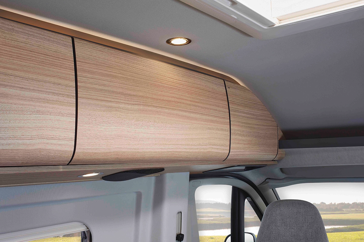 Armadietti in legno con finitura liscia a venature chiare e luci LED integrate all'interno di un moderno abitacolo di camper, sopra il sedile del conducente e il finestrino.