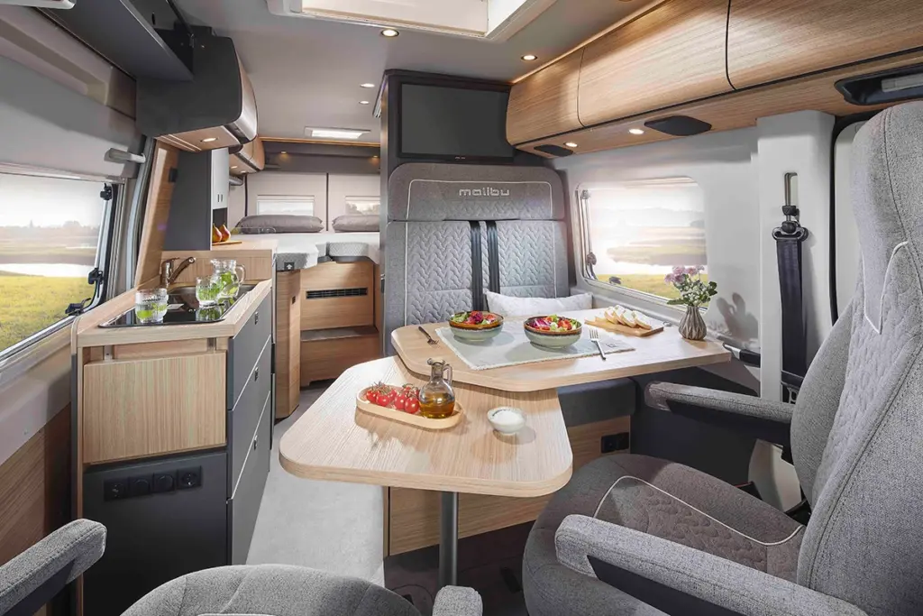 Modern camperinterieur met grijze zetels, een eettafel met salade, brood en drankjes, een compacte keuken met verse groenten en een knus bed op de achtergrond. Natuurlijk licht stroomt binnen door grote ramen en een dakraam.