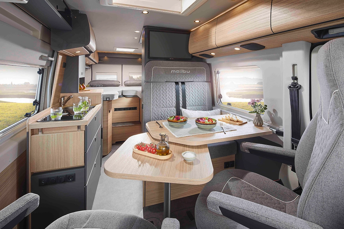 Interno di un moderno camper con tavolo da pranzo apparecchiato con insalata, pane e bevande. Lo spazio è caratterizzato da sedili grigi, armadietti in legno, un piccolo angolo cottura e finestre che lasciano entrare la luce naturale.