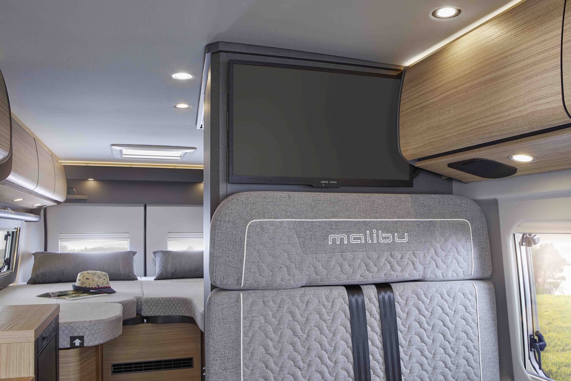 L'interno di un moderno camper con sedile grigio Malibu, TV a schermo piatto, mobili in legno e un accogliente letto posteriore con cuscini e cappello sul letto.