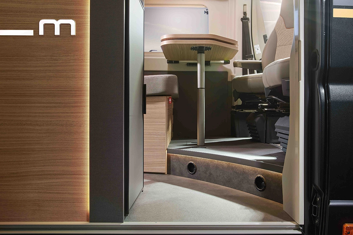 Vista dalla porta aperta di un moderno camper, che mostra un gradino che porta a una zona pranzo compatta con tavolo in legno e sedili beige, e luci soffuse lungo la base dell'interno.