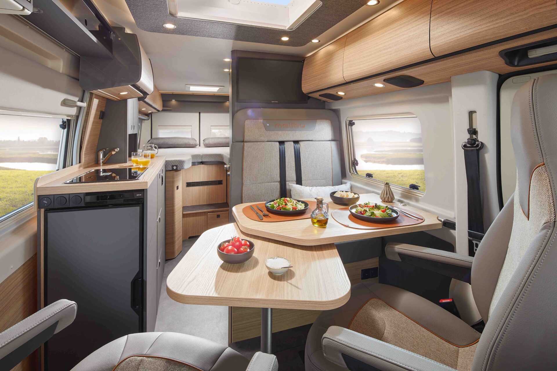 Modernes Wohnmobil-Interieur mit einem Holztisch für eine Mahlzeit, zwei Drehstühlen, einer kompakten Küche und großen Fenstern, die natürliches Licht hereinlassen. Der Tisch bietet Platz für Teller und Gläser und schafft einen gemütlichen Essplatz.