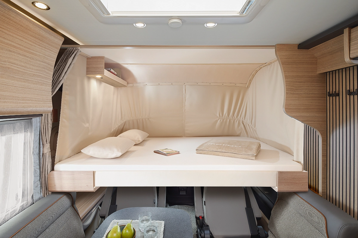 Ein kompaktes Wohnmobil-Interieur mit einem erhöhten Bett mit beigem Bettzeug, Kissen und einem Buch darauf. Unter dem Bett befindet sich ein kleiner Essbereich mit einem Tisch, auf dem Gläser, ein Krug und grüne Birnen stehen. Holzverkleidung schmückt den Raum.