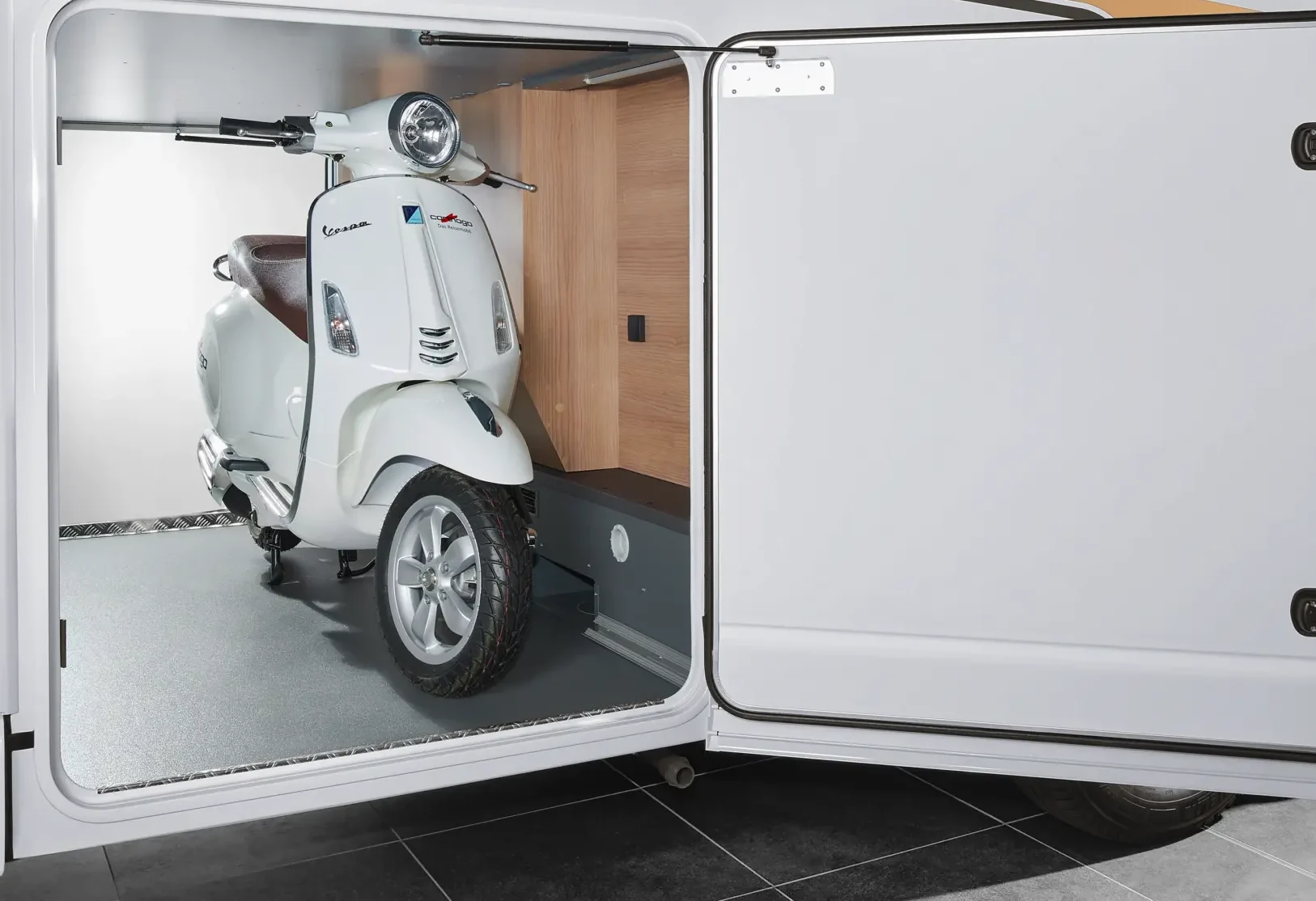 Ein weißer Motorroller ist im Stauraum eines weißen Malibu-Wohnmobils geparkt, wobei die Seitentür geöffnet ist und der Roller ordentlich gesichert im Inneren liegt.