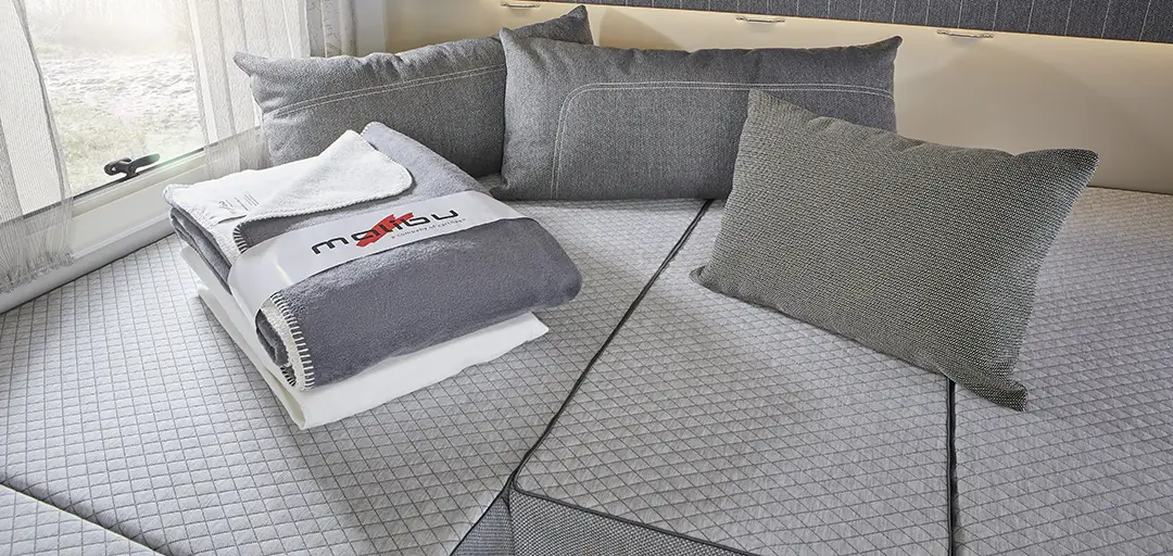 Ein gemütliches Wohnmobilbett mit grauen Kissen und Polstern, ordentlich gefalteten Handtüchern und einer Decke obenauf, unter Dachschränken und neben Fenstern mit durchsichtigen Vorhängen.