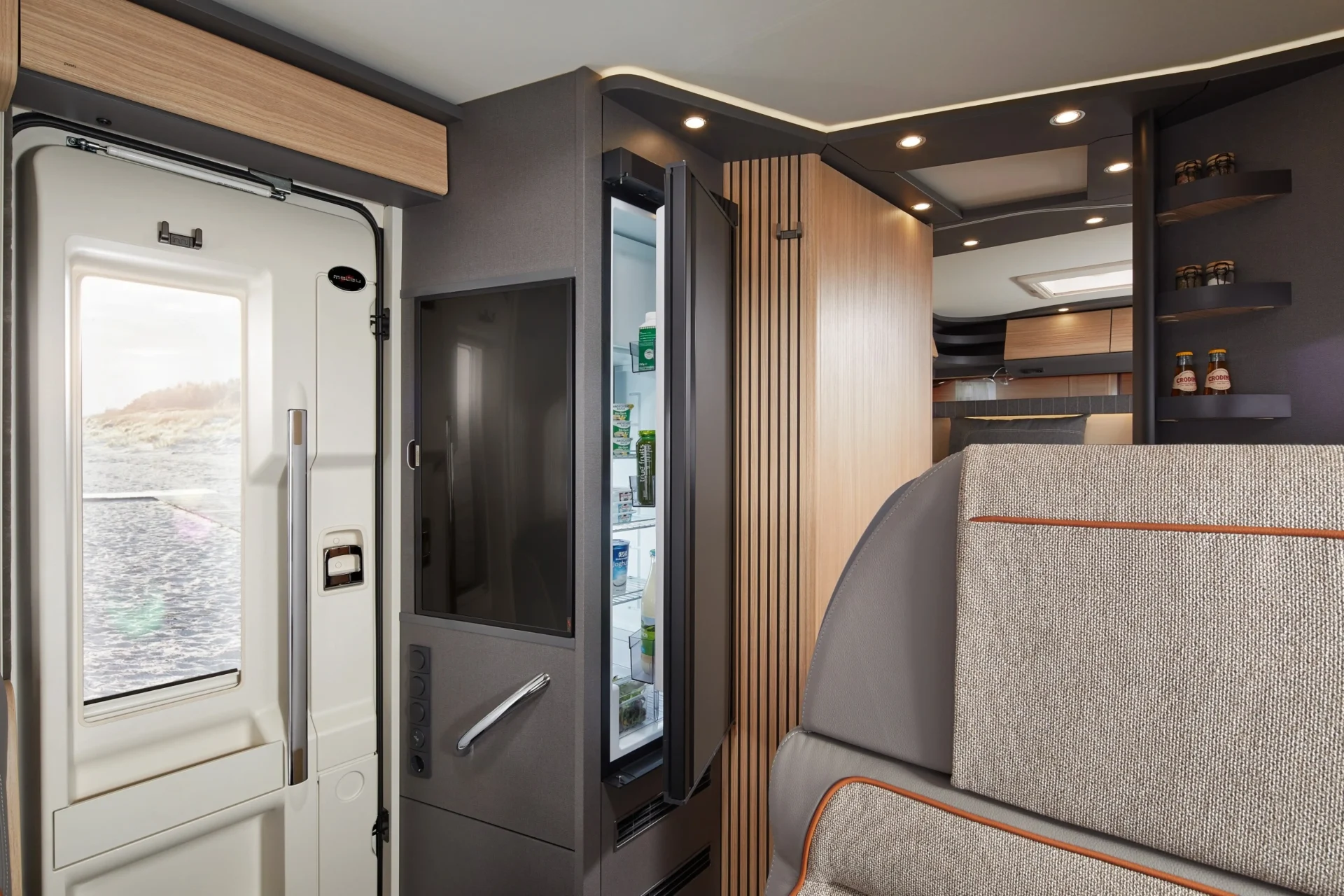 Modernes Wohnmobil-Interieur mit einem teilweise offenen Kühlschrank, Regalen mit Gewürzen, einem gepolsterten Sitz und einer Tür nach draußen mit Blick auf einen Strand. Der Raum wirkt hell, organisiert und komfortabel.